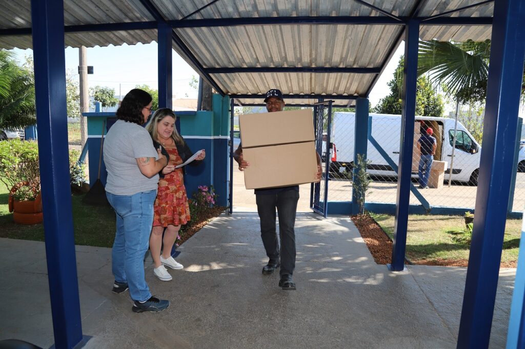 Diretora da escola conferindo a entrega de uniformes enquanto homens descarregam o caminhão com as caixas