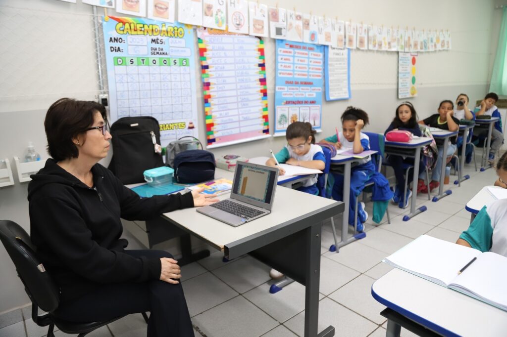 Professora sentada em frente ao seu chromebook, em sala de aula.