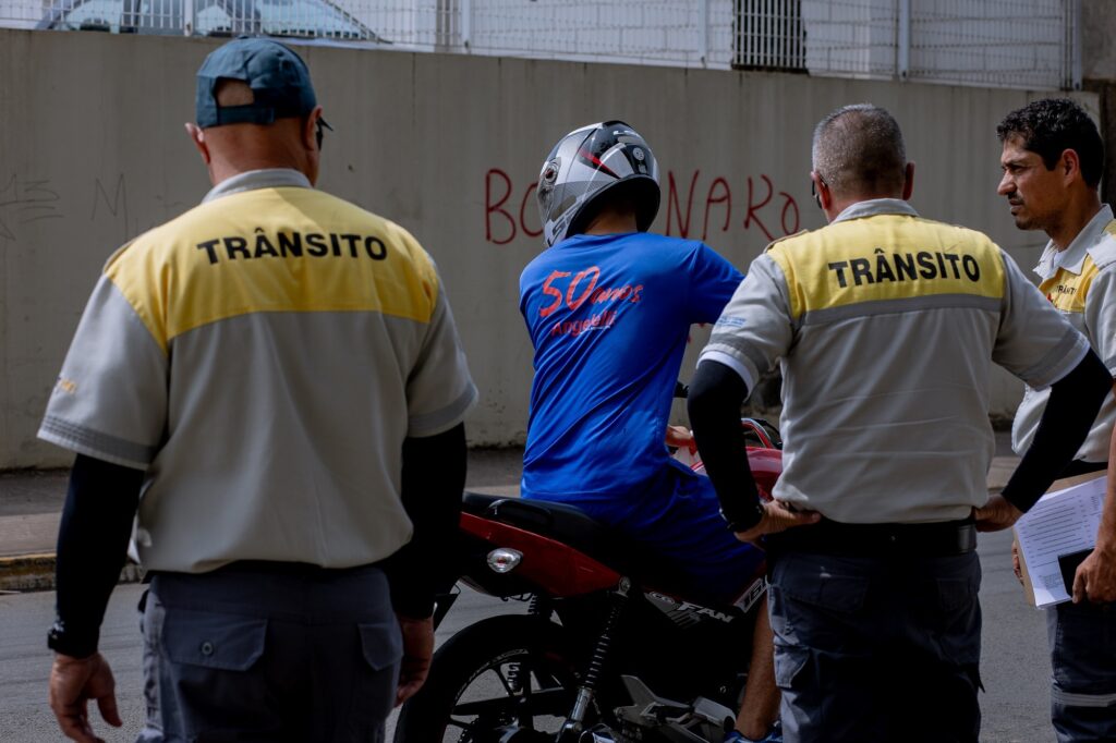 Três agentes de trânsito com uniforme cinza e amarelo, observando um motoqueiro de camiseta azul, em cima da moto vermelha e preta