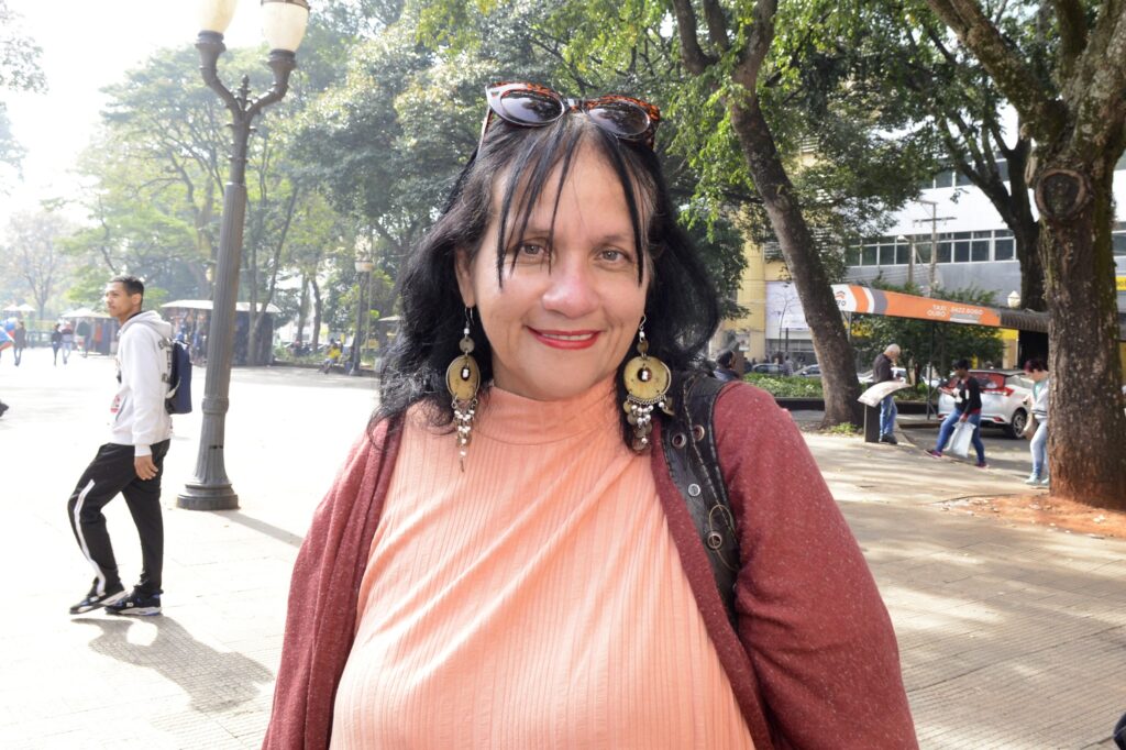 Mulher de cabelos escuros na altura do ombro, com brincos grandes, óculos escuros na cabeça, camiseta laranja e casaco laranja escuro, com paisagem de fundo de praça