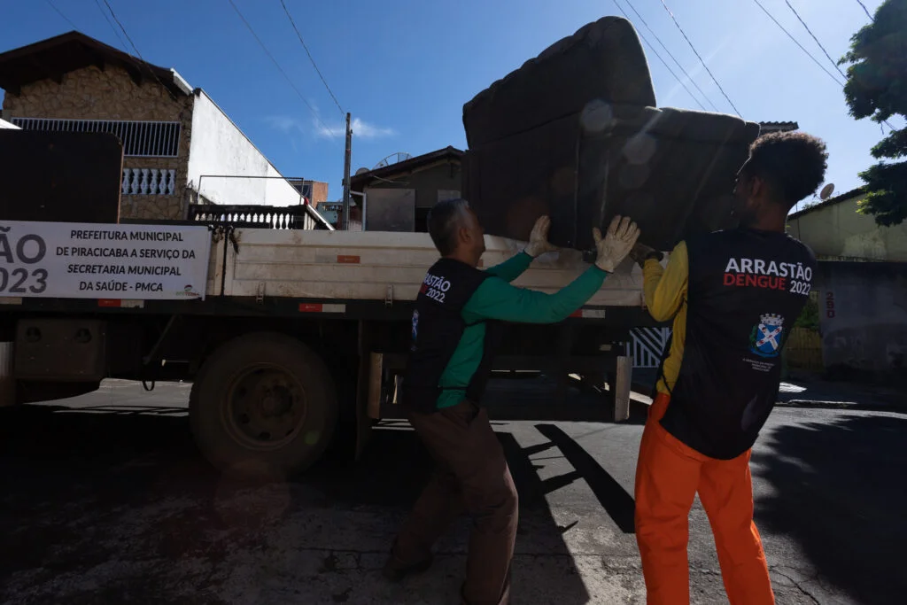 Imagem mostra funcionário da prefeitura com uniforme da cor laranja e colete azul escuro ao lado de uma caminhão onde ele deposita materiais inservíveis, como cadeiras e sofás