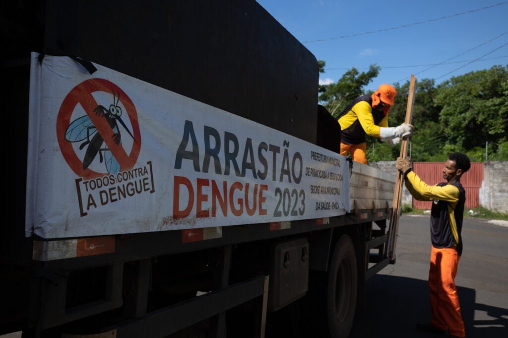 Imagem mostra uma homem coletando inservíveis na rua e entregando para outro que está em cima de um caminhão com adesivo que contém os seguintes dizeres: Arrastão da Dengue 2023