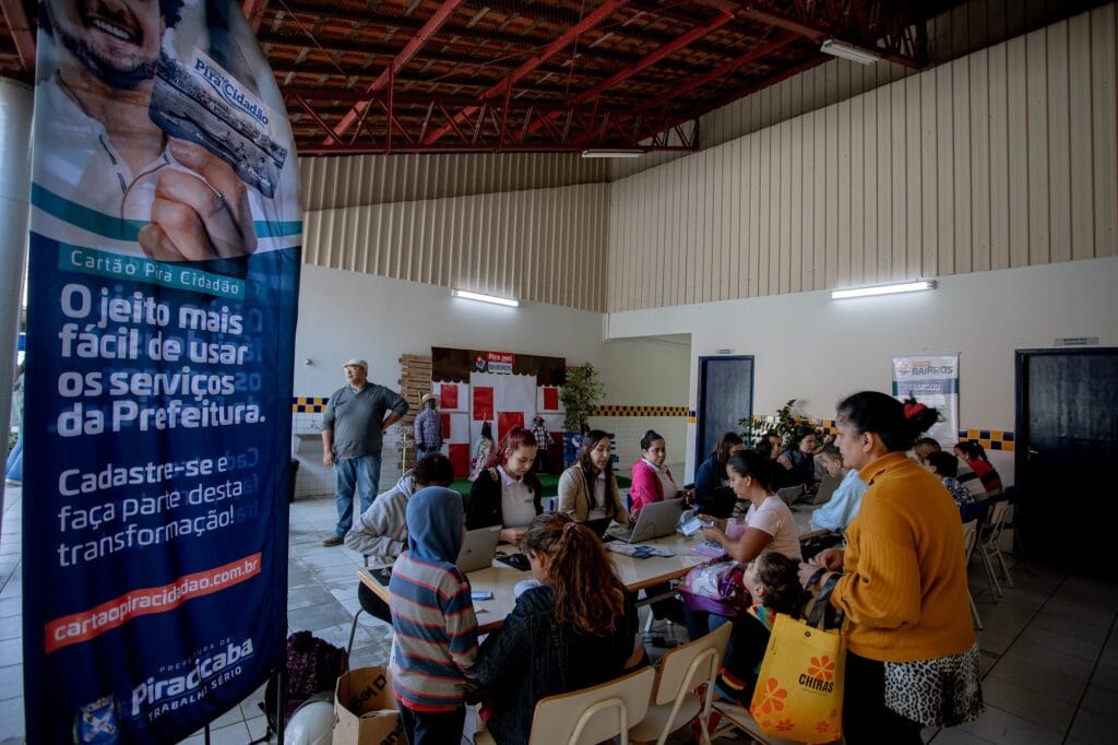 Diversas pessoas sendo atendidas em uma mesa e destaque para um banner, no canto esquerdo da imagem, que tem informações sobre o cartão Pira Cidadão.