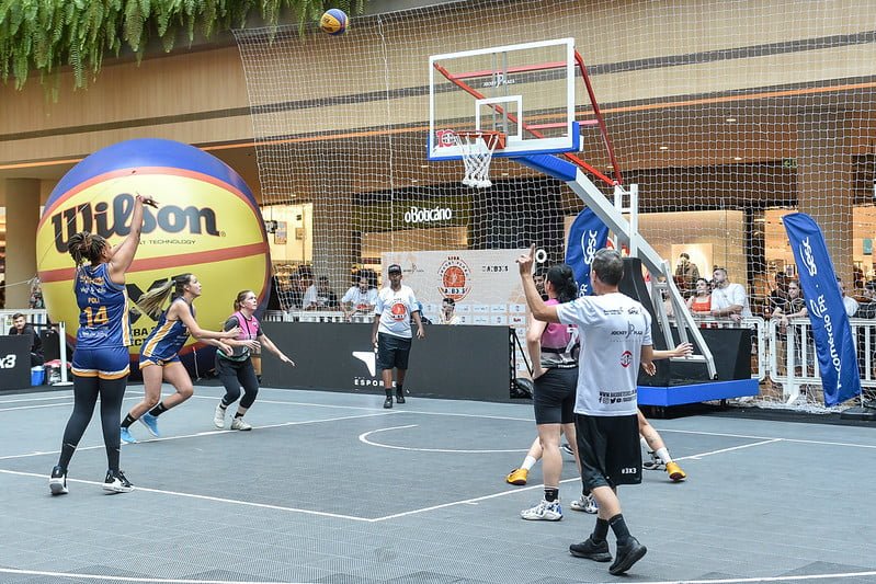 Atletas de basquete, da modalidade 3x3 disputam uma partida.
Uma menina com calção e camiseta azul, observada por suas companheiras e adversárias, faz o arremesso.