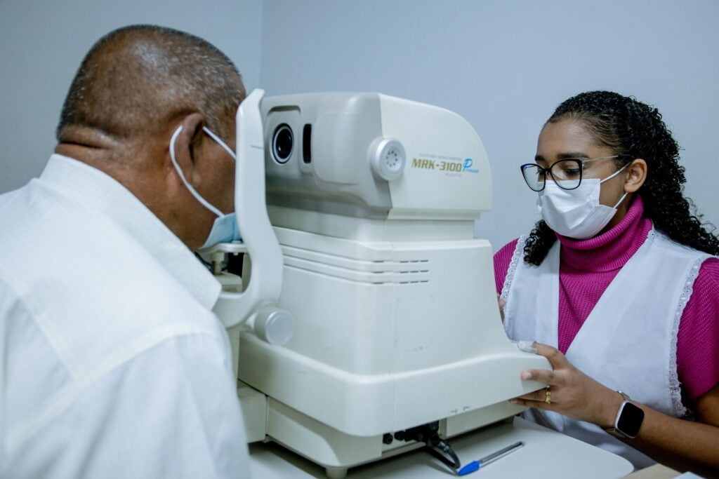 Imagem colorida mostra paciente colocando o rosto em máquina para realizar exame ocular e, ao seu lado, enfermeiro acompanha todo o procedimento