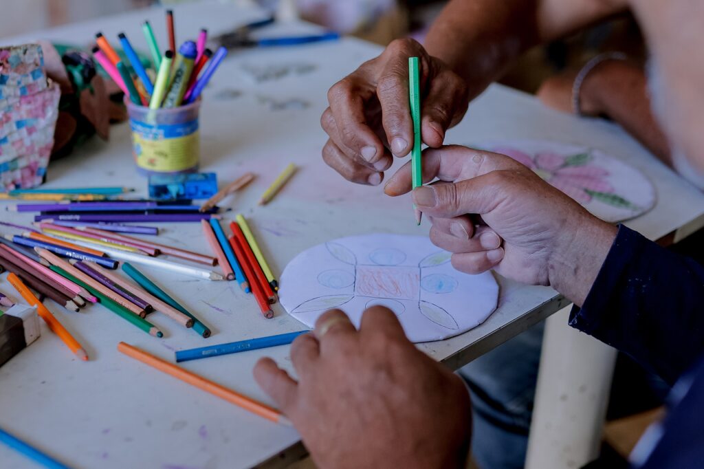 em uma mesa branca, lápis e pinceis de várias cores são utilizados pelos moradores para artesanato e criação de mosáicos