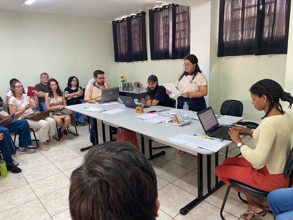 Na imagem, Claudia Novolette, presidente do Comsea em pé, atrás da mesa e candidatos à frente.
