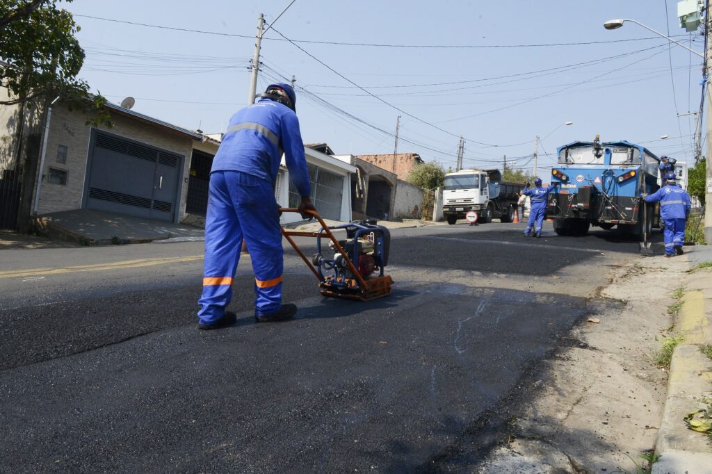 Imagem de um funcionário em roupa azul atuando com compactador no asfalto durante os serviços de tapa-buraco. Ao fundo, estão dois caminhões.