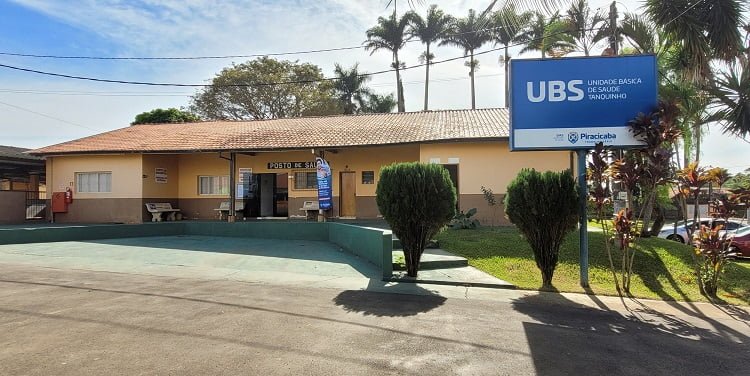 imagem colorida mostra a frente da unidade de saúde Tanquinho com sua grande escadaria e jardim com flores, coqueiros e grandes pinheiros