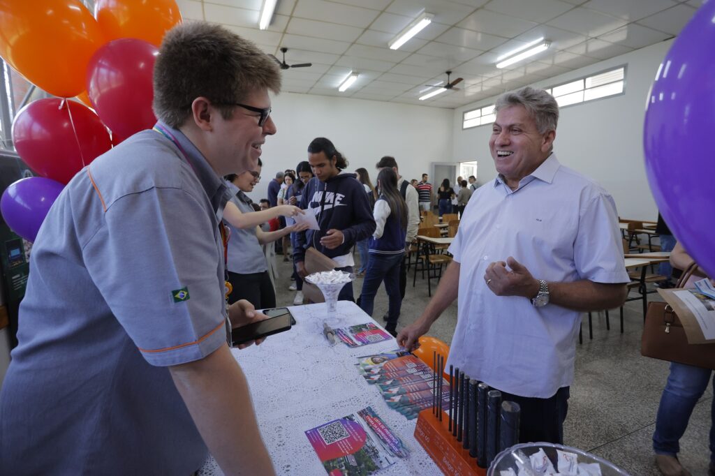 Imagem do secretário José Luiz Ribeiro (à direita) conversando com um homem. A imagem também mostra pessoas ao fundo, em fila, sendo atendidas