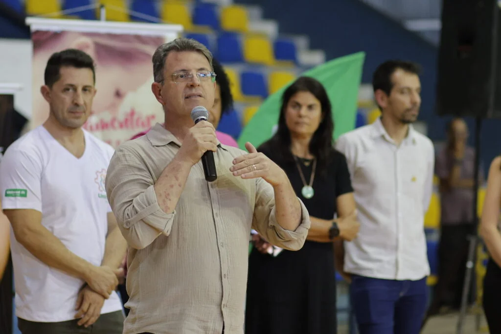Em evento no ginásio municipal de esporte, o médico Rogário Tuon usa microfone para conversar com as mães presentes em evento do Mamaço