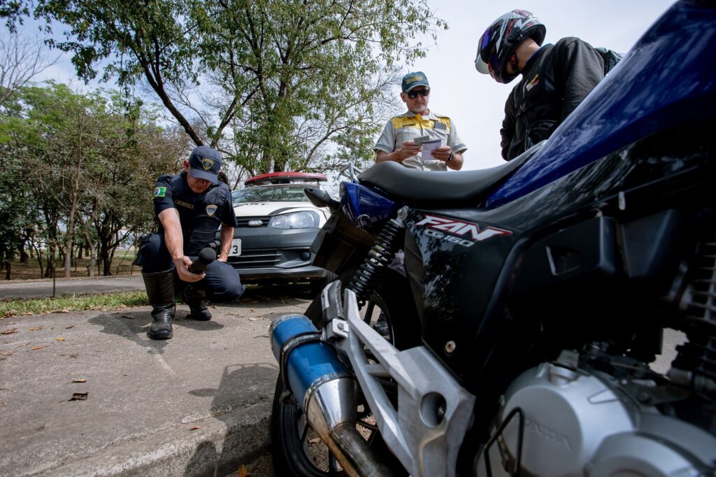 Guarda Civil de uniforme azul escuro abaixado, tendo ao lado um agente de trânsito de uniforme cinza e amarelo, conferindo a documentação de motoqueiro de roupa preta