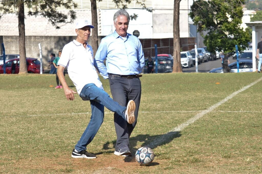 Homem de boné chuta a bola, observado pelo secretário de Esportes, Hermes Balbino, que está de camisa longa azul