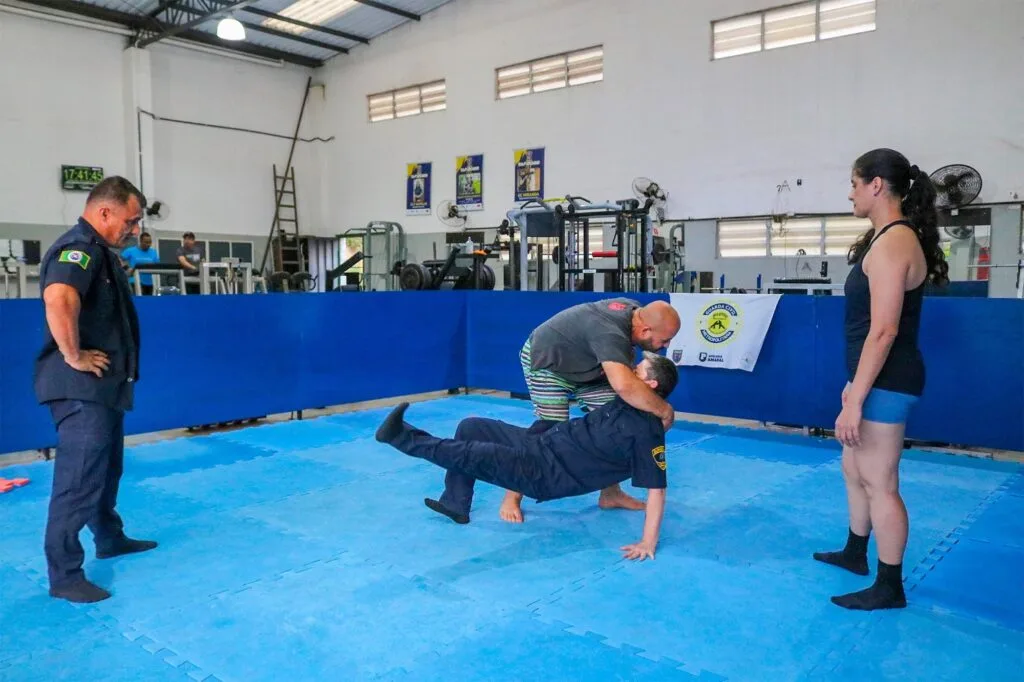 Professor de camiseta cinza, segura guarda civil, de uniforme escuro que está caindo, observado por um guarda homem e outra guarda mulher