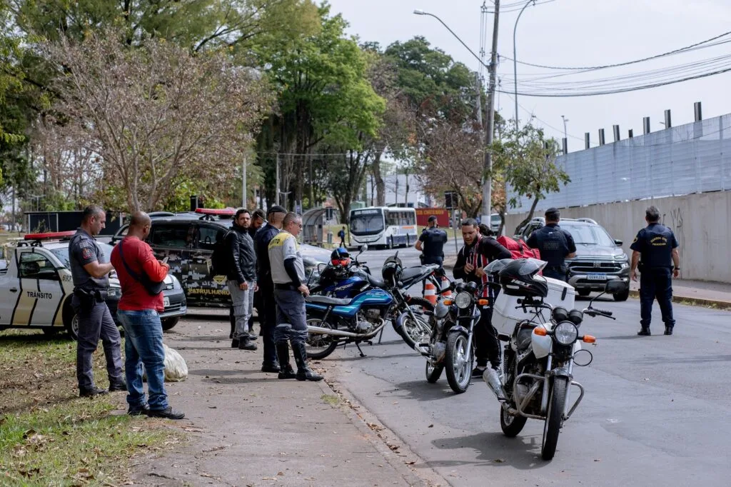 Motoqueiro de camisa listrada vermelho e preto, para para sua moto ser vistoriada, observada por agente de trânsito de bota e uniforme cinza e amarelo