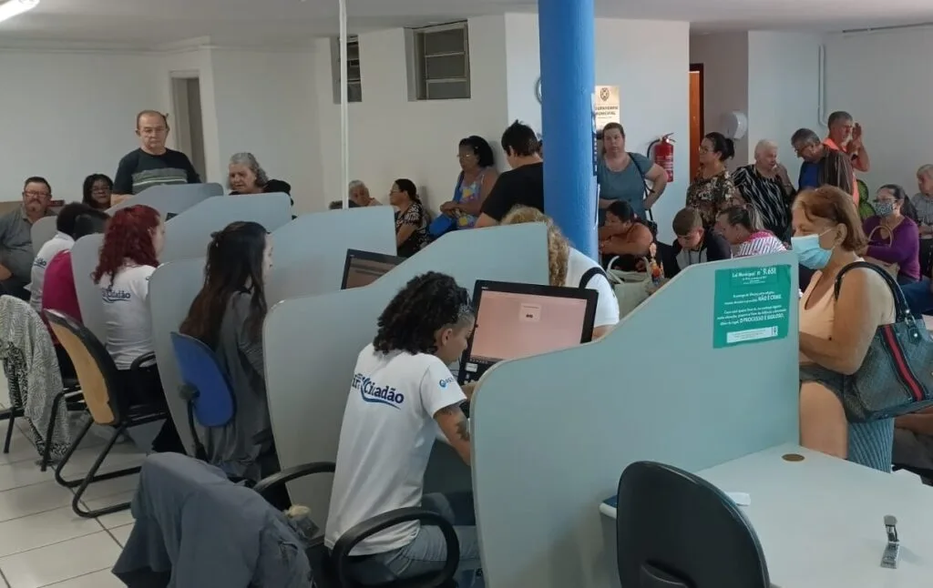 Dezenas de pessoas fazem fila dentro do poupatempo municipal para realizarem seu cadastro com as quatro atendentes uniformizadas