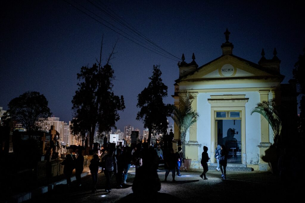 Imagem do cemitério à noite, com prédio ao fundo e pessoas ao redor, em ambiente noturno