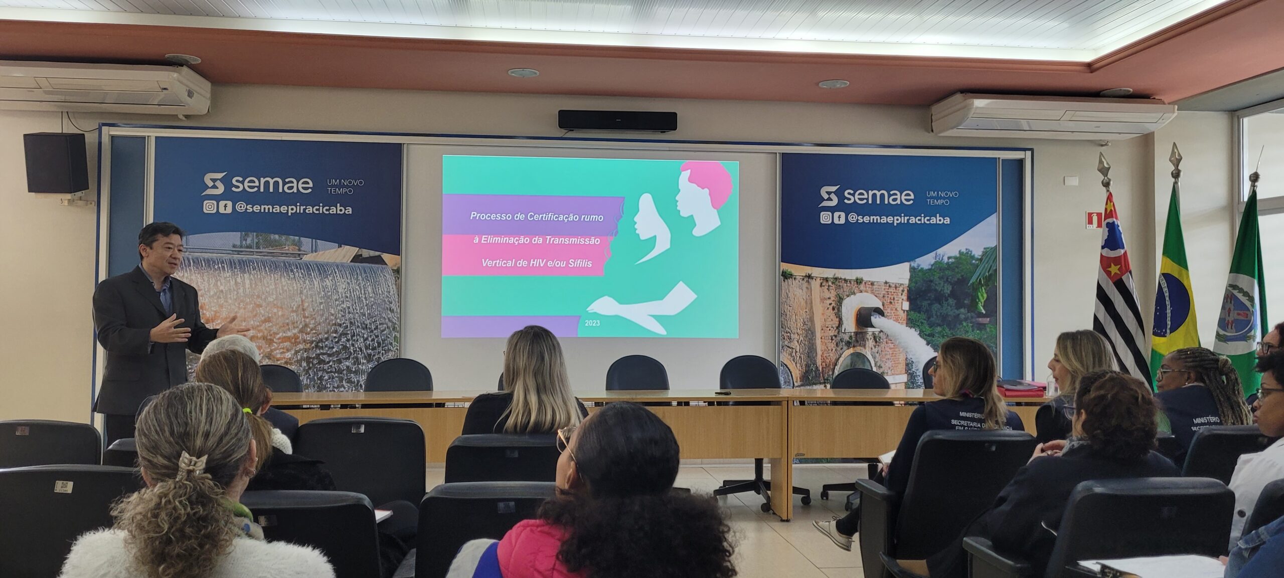 Secretário Douglas Koga está em pé na frente do público no auditório do Museu da água. Ao fundo no telão colorido, as informações sobre a transmissão vertical de HIV na cidade