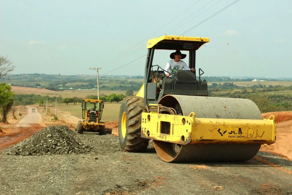 Rolo compactador da Sema compactando o solo e motoniveladora ao fundo espalhando o material lajão britado