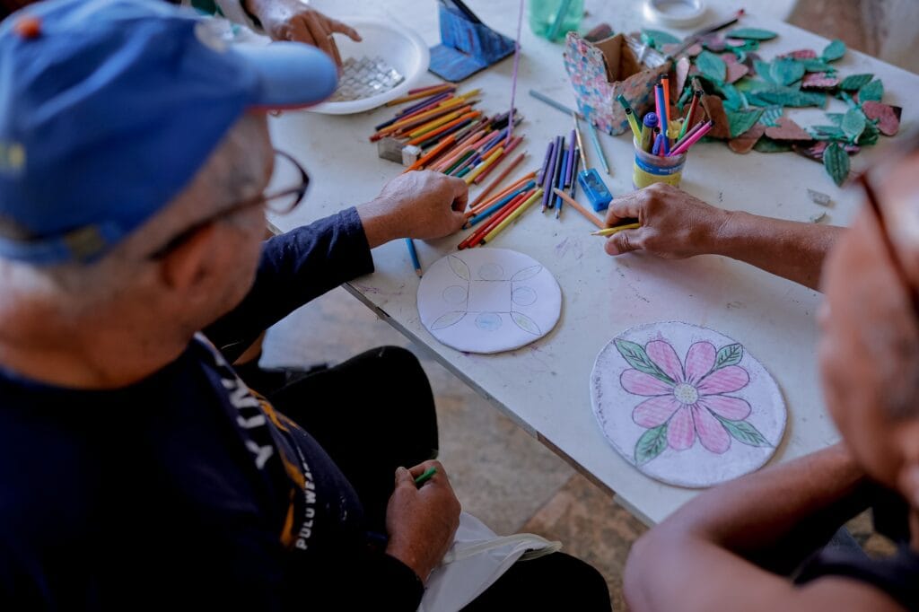 com boné azul J.L. faz pintura e cria mosaico com azulejo e tintas de várias cores que estão sobre uma mesa branca