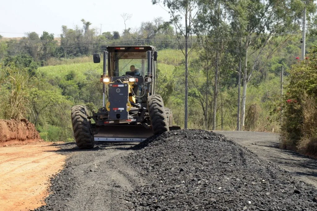 Imagem de uma máquina aplicando as raspas de asfalto na estrada; tem árvores ao fundo