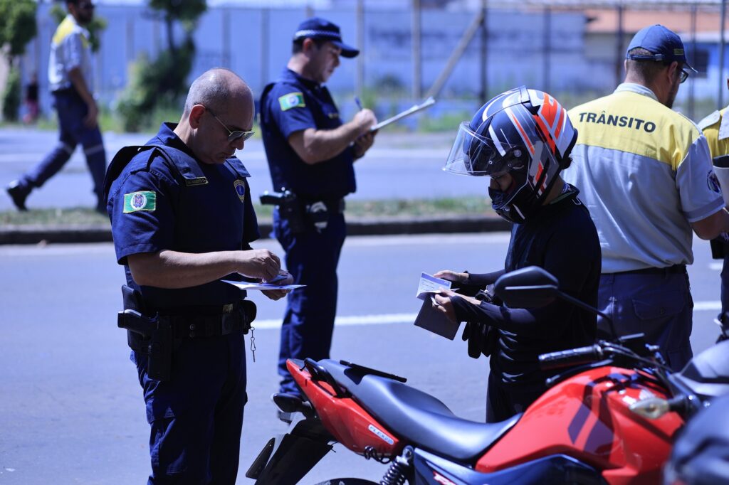 homens em pé e outro com capacete na rua, ao lado de moto