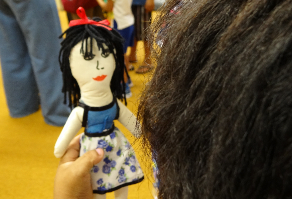 Na imagem, mulher de costas segura boneca de pano que confeccionou. A boneca é branca, com cabelo preto, laço vermelho na cabeça e roupa azul e florida