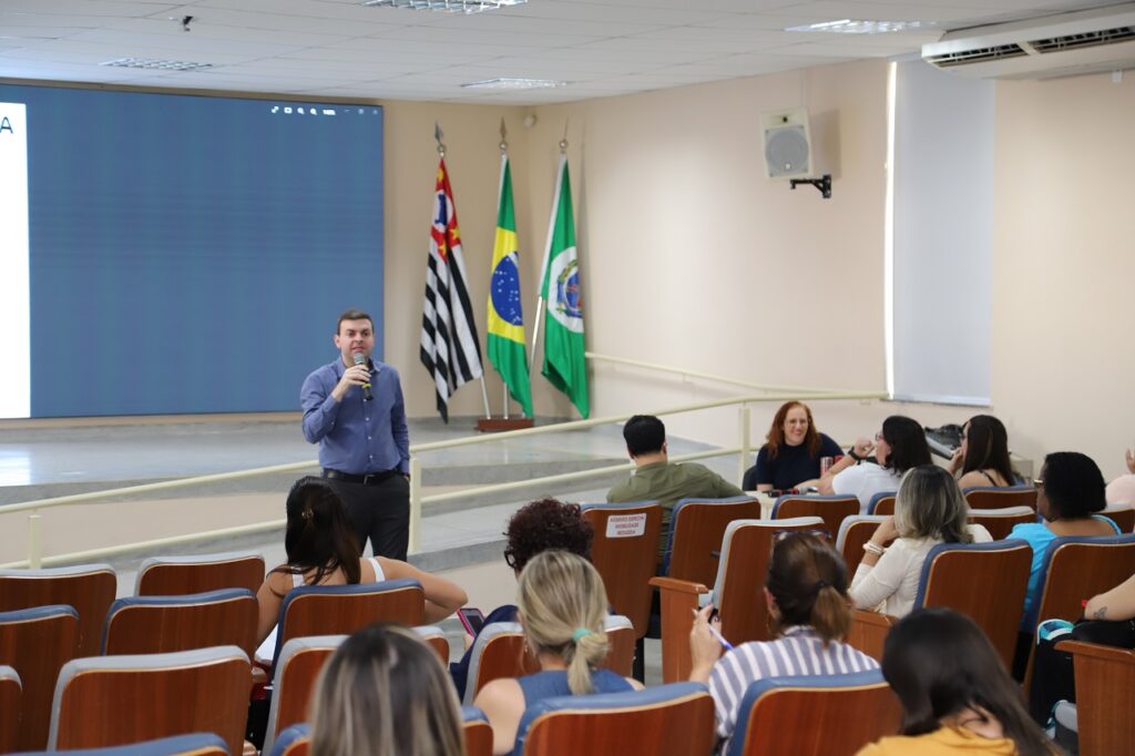 Secretário de Educação, Bruno Roza, em pé no anfiteatro da Secretaria Municipal de Educação, falando ao microfone durante a reunião com os diretores das escolas.