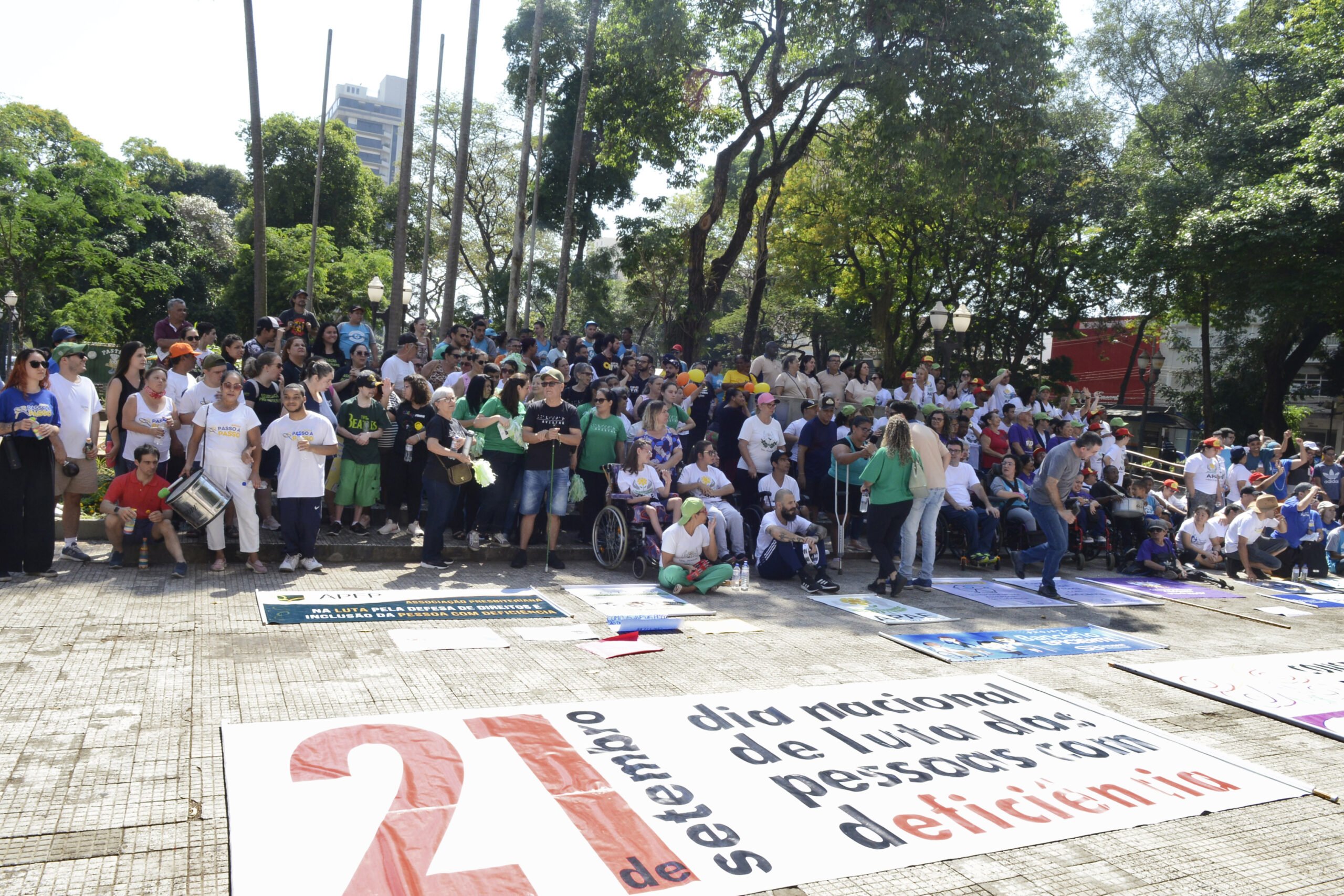 Ao fundo da imagem, participantes da caminhada da inclusão em pé atrás da faixa 21 de setembro - Dia Nacional de luta da Pessoa com Deficiência