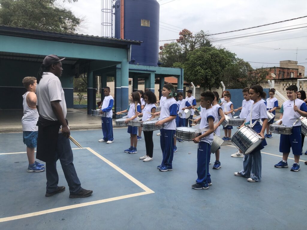 Alunos na quadra da escola com instrumentos da fanfarra, vestidos todos de uniforme, acompanhando as instruções do professor, durante ensaio para o desfile.