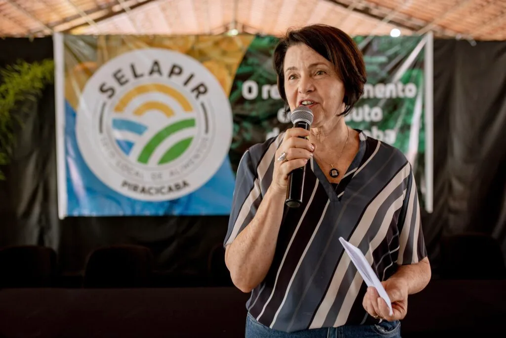 Secretária Nancy Thame discursando com um microfone, durante evento de lançamento do Selo Local de Alimentos de Piracicaba, em novembro de 2022