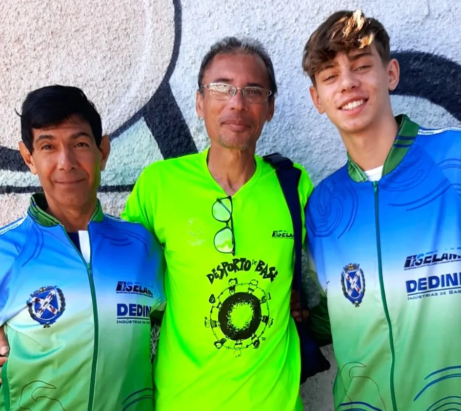 Professor de atletismo veste roupa verde, abraçado de dois atletas, um senhor de 60 anos e outro de 15 anos, que vestem uniforme azul da secretaria de esportes de Piracicaba.