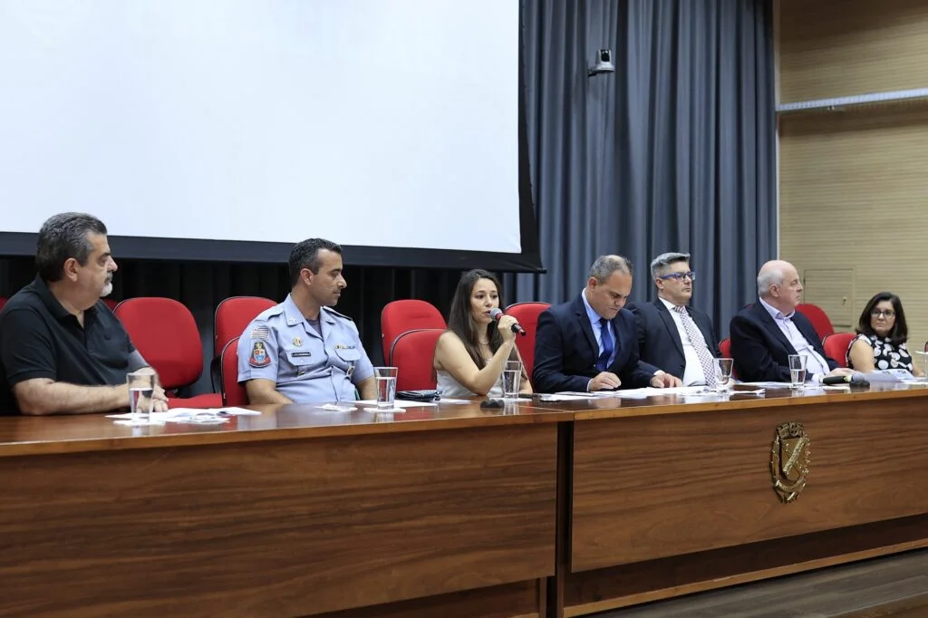Autoridades sentadas a mesa e a secretária de Trânsito, Jane Franco, ao centro segurando o microfone