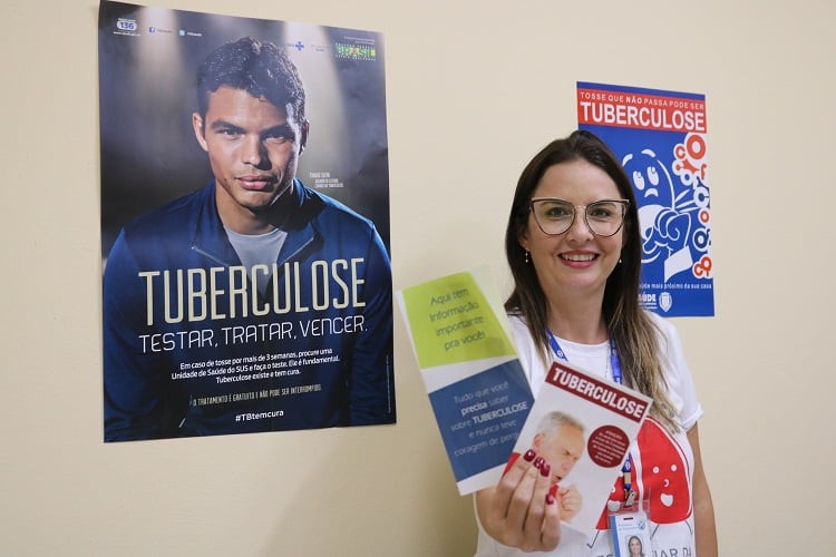 Poliana Garcia exibe um dos folhetos educativos que são entregues a população sobre tuberculose; ao fundo, na parede, está evidenciado dois cartazes com dizeres sobre a prevenção da doença