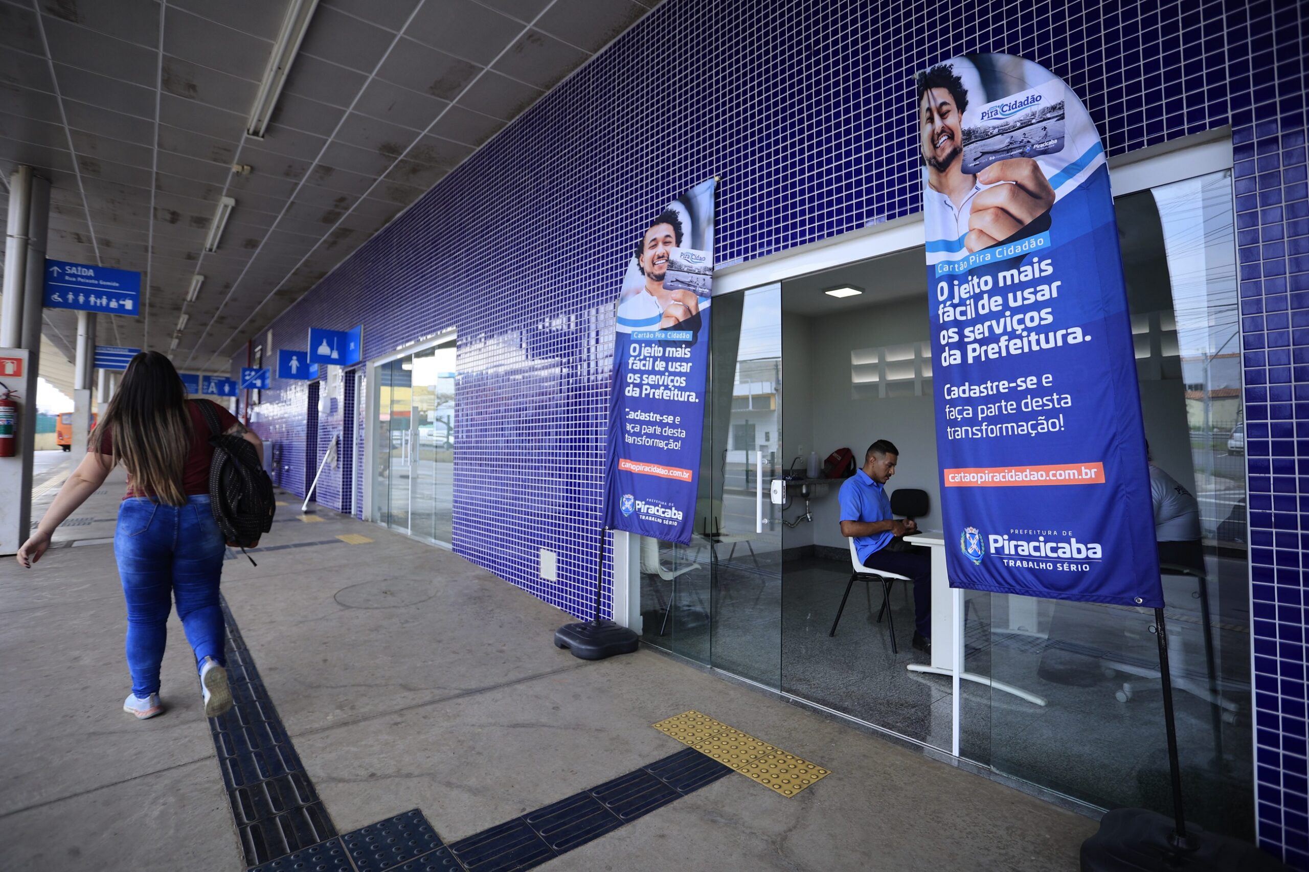 Pessoas caminham pelo terminal de ônibus do Vila Sônia. em uma das salas, de paredes azuis e identificada com bandeiras na frente, equipe do cartão Pira Cidadão faz o credenciamento da população