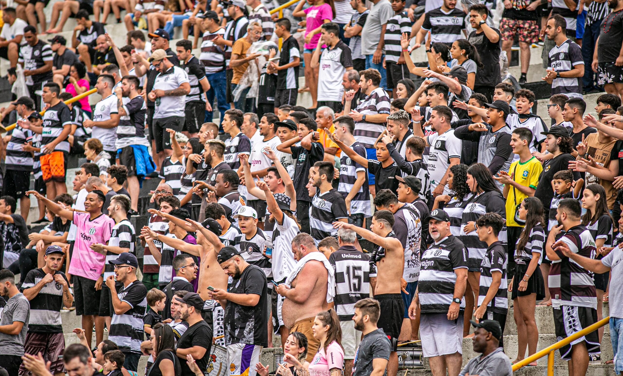 Imagem colorida mostra a grande torcida do XV de Piracicaba dentro do estádio Barão da Serra Negra. As pessoas estão vestidas com a camisa do alvinegro e agitam as mãos para incentivar o time em campo