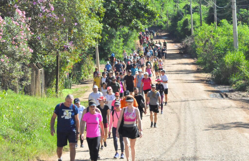 Pessoas estão caminhando por uma estrada de terra na região de Piracicaba.A atividade faz parte do Caipirandando, promovida pela Secretaria de Esportes de Piracicaba.Na foto é possível ver várias pessoas caminhando, numa área rural com roupas coloridas, bermudas, bonés.