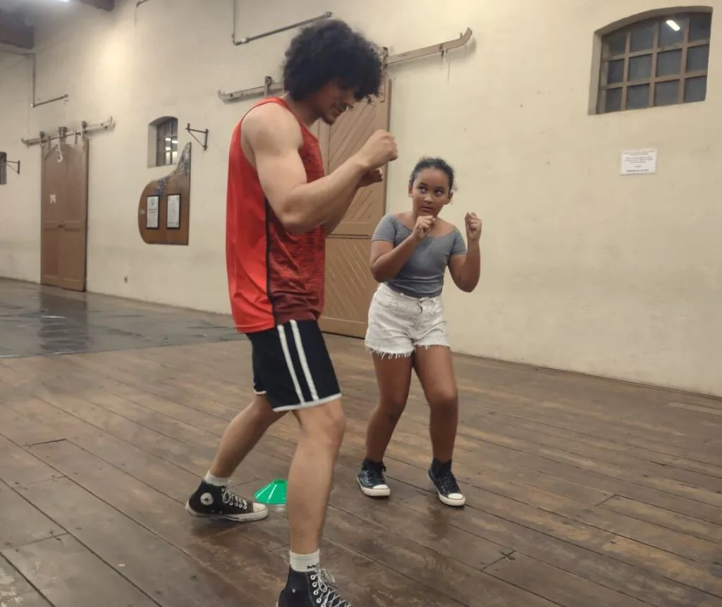Uma menina de 9 anos, vestindo blusa cinza e calção branco, com tênis preto, observa os movimentos do professor de boxe, que veste roupa vermelha e preto.