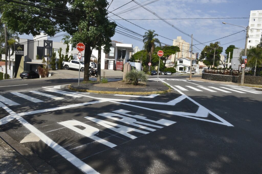 Faixas brancas e a letra PARE na cor branca, no asfalto de avenida.