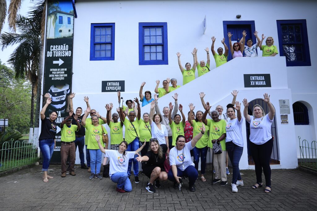 Homens e mulheres adultos com os braços levantados, representando alegria, em frente ao Casarão do Turismo, prédio de antiga olaria de Piracicaba