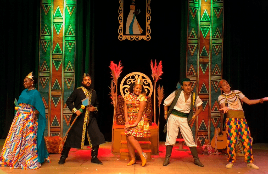 Imagem de um cenário de para teatro, com duas colunas com imagens geométricas, um trono dourado no meio, com uma mulher com coroa sentada, com um homem e uma mulher de um lado e dois homens do outro.