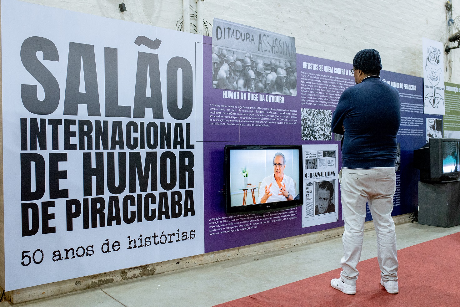 Na imagem, homem observa painel ao fundo com o texto Salão Internacional de Humor de Piracicaba e retrospectiva com imagens.