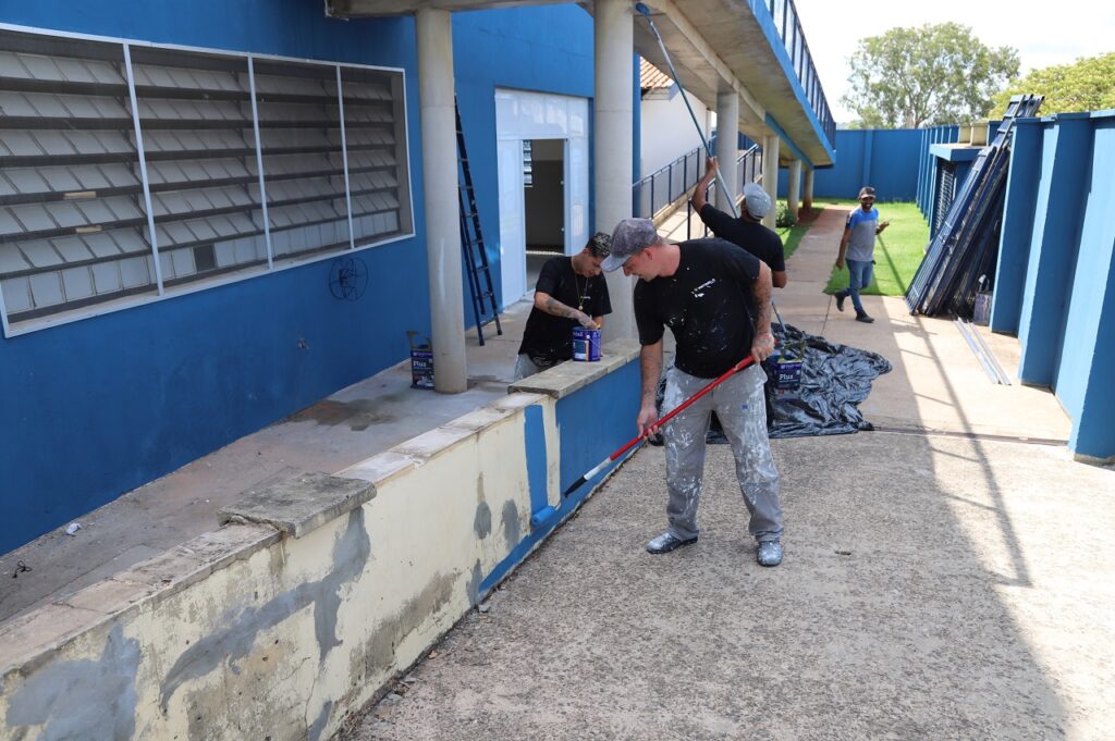 a imagem mostra 4 homens que estão pintando a parte de fora da escola. Os homens estão uniformizados com rolos de tinta pintando uma passarela e muretas da rampa de acesso ao prédio.