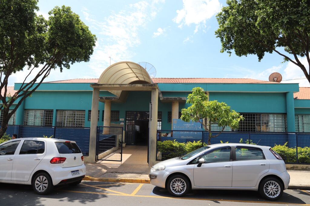 A imagem mostra a frente da escola após a reforma, pintada com as cores da prefeitura. Tem dois carros parados em frente a escola que aparecem na imagem.