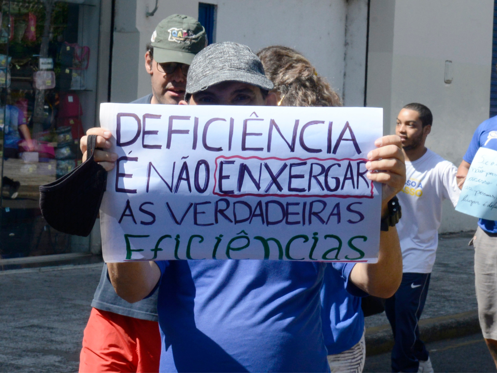 Na imagem, menino esconde o rosto com cartaz que diz: Deficiência é não enxergar as verdadeiras eficiências