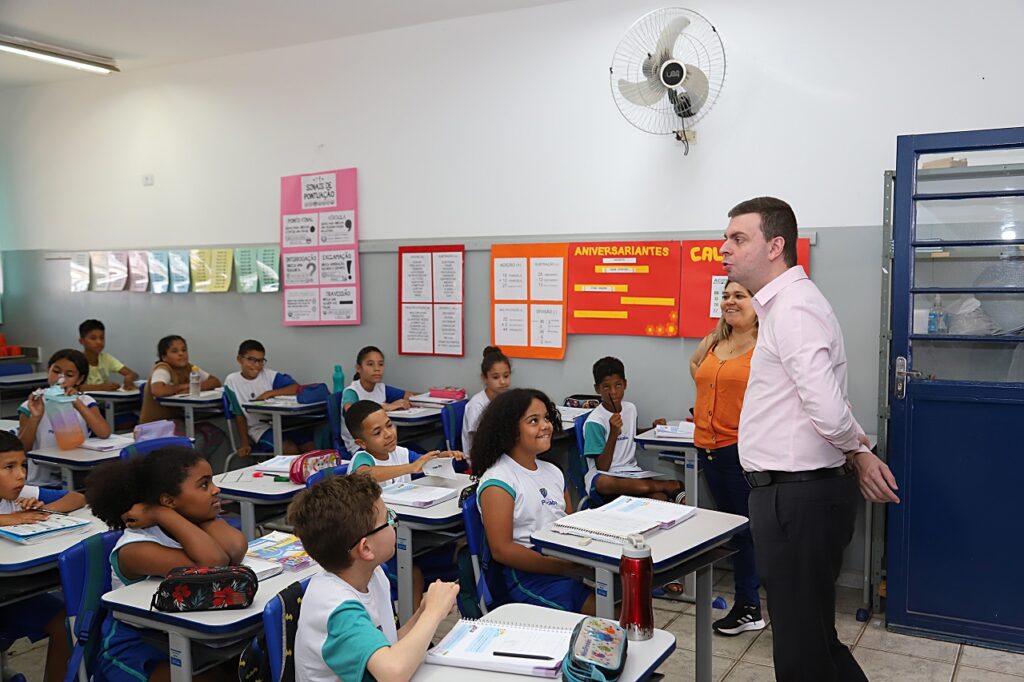 A imagem mostra o secretário Bruno Roza em sala de aula, em pé conversando com os alunos que estão sentados e uniformizados, atentos e prestando atenção na conversa.