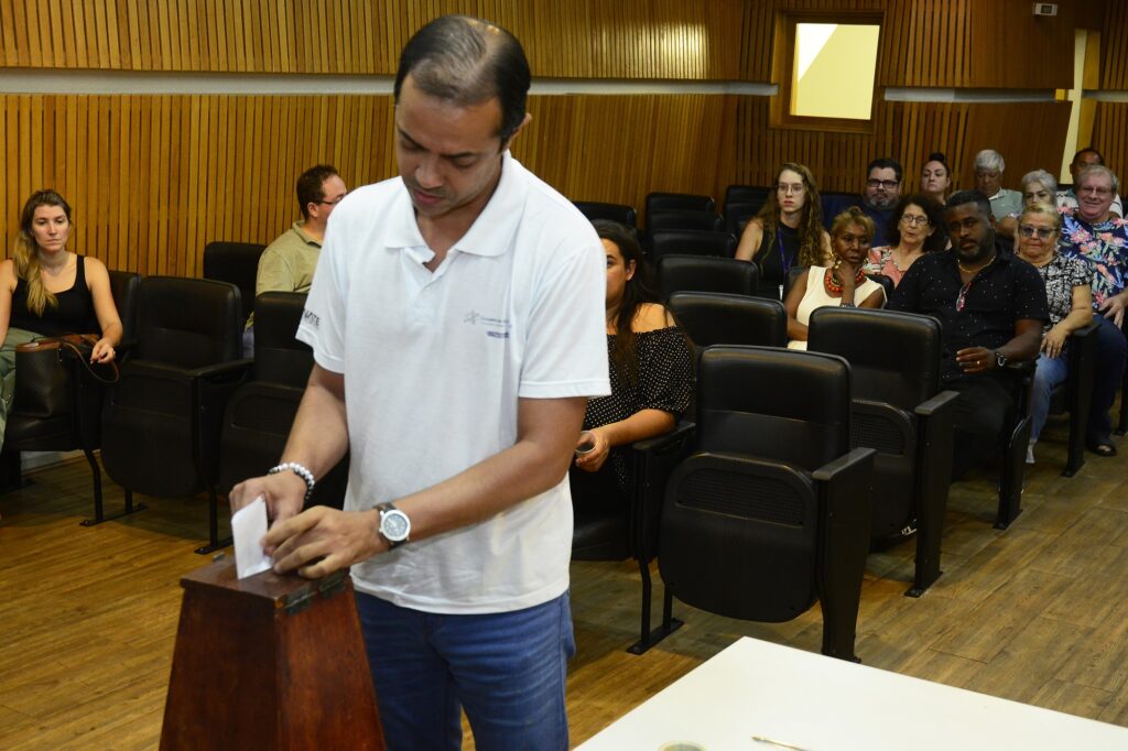 Rapaz de camiseta branca e calça jeans, deposita seu voto na eleição do Conselho Municipal do Idoso.