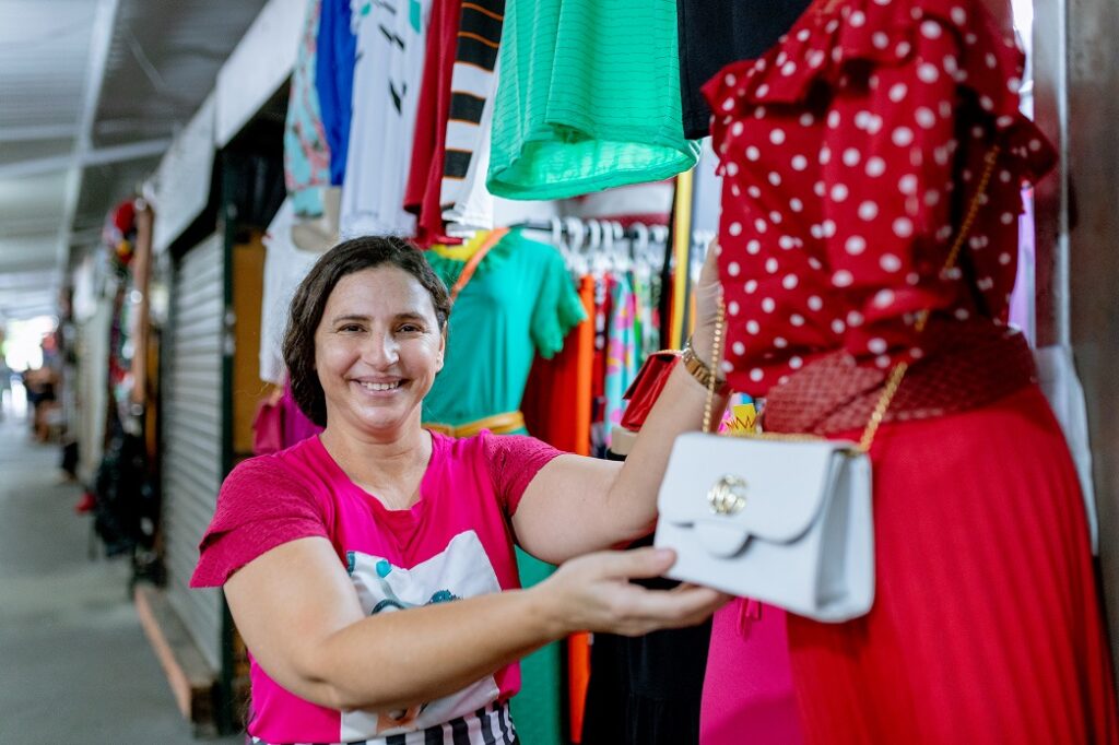 Mulher sorrindo segura bolsa branca com as mãos, dependurada em banca junto com diversas peças de roupa ao lado e ao fundo