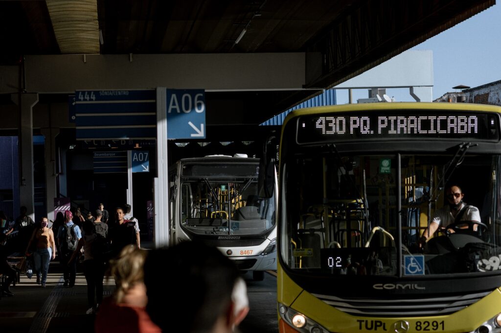 imagem de dois ônibus dentro de terminal de ônibus com pessoas ao redor