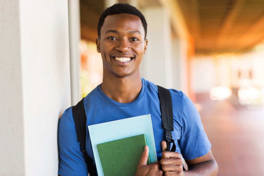 Jovem negro sorridente de camiseta azul, segurando material escolar.